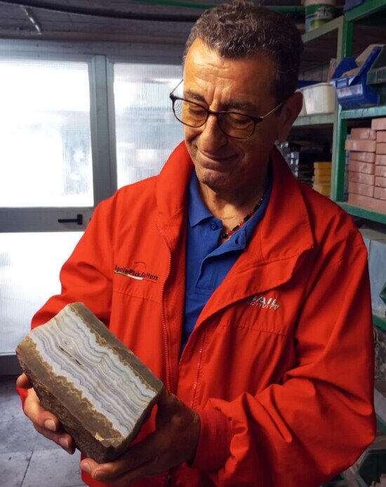 In immagine il titolare di agatepick nel magazzino dell'azienda con in mano un pesante blocco di pietra di agata. Vestito con un giubbino estivo rosso vivo e con occhiali da vista mentre con un sorriso guarda il blocco di pietra con ammirazione