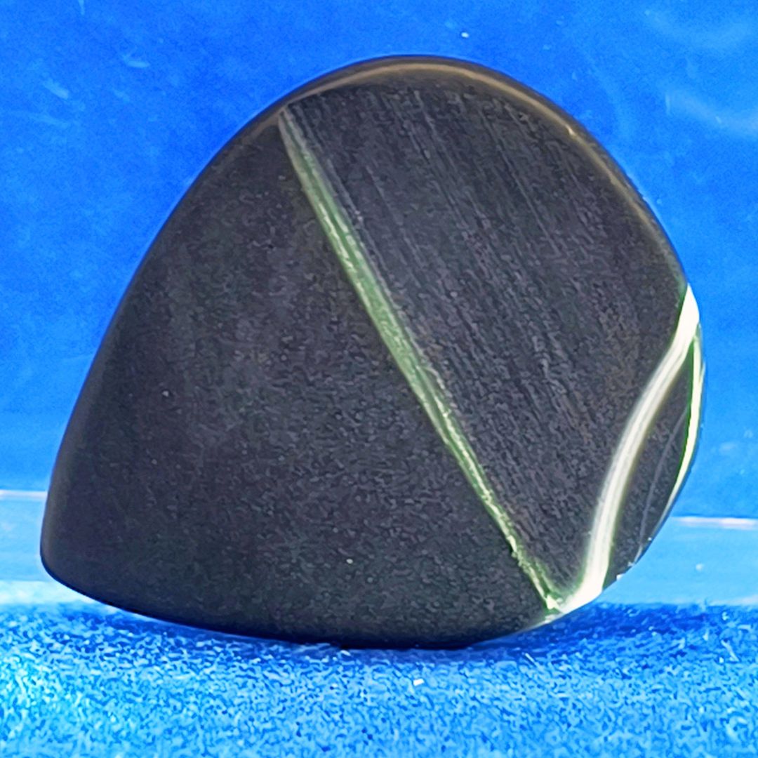 Plettro in pietra di onice nera con striature bianche trasparenti e adesivo con micro granuli di quarzo per ottimizzare il grip e la presa sul codolo del plettro, fotografato con sfondo azzurro.