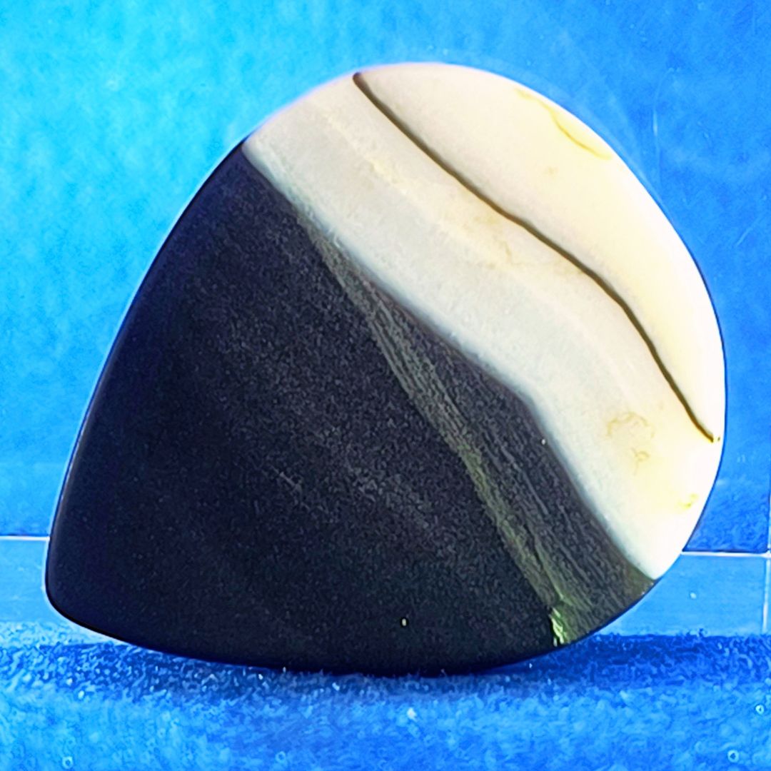 Plettro in pietra di onice nera con striature bianche trasparenti e adesivo con micro granuli di quarzo per ottimizzare il grip e la presa sul codolo del plettro, fotografato con sfondo azzurro.